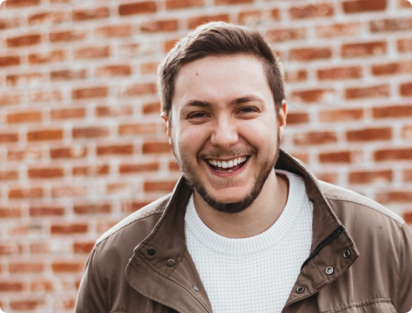 Young man in brown jacket grinning in front of brick wall
